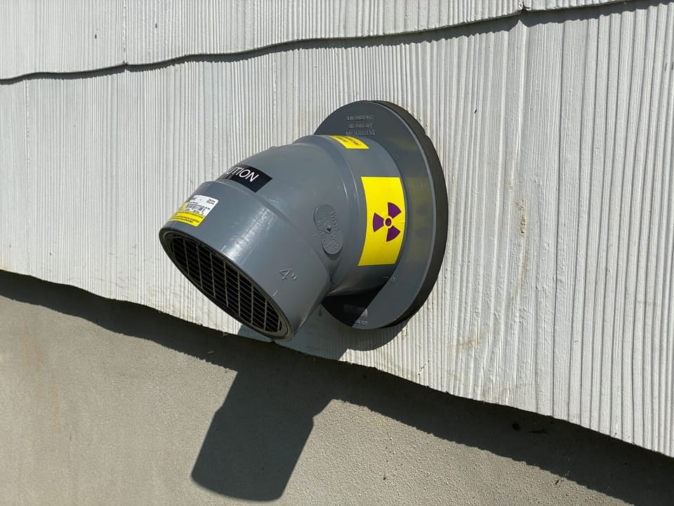 A grey four inch radon exhaust vent on the exterior wall of a house, carrying the yellow radiation trefoil label that identifies it as part of a radon system.